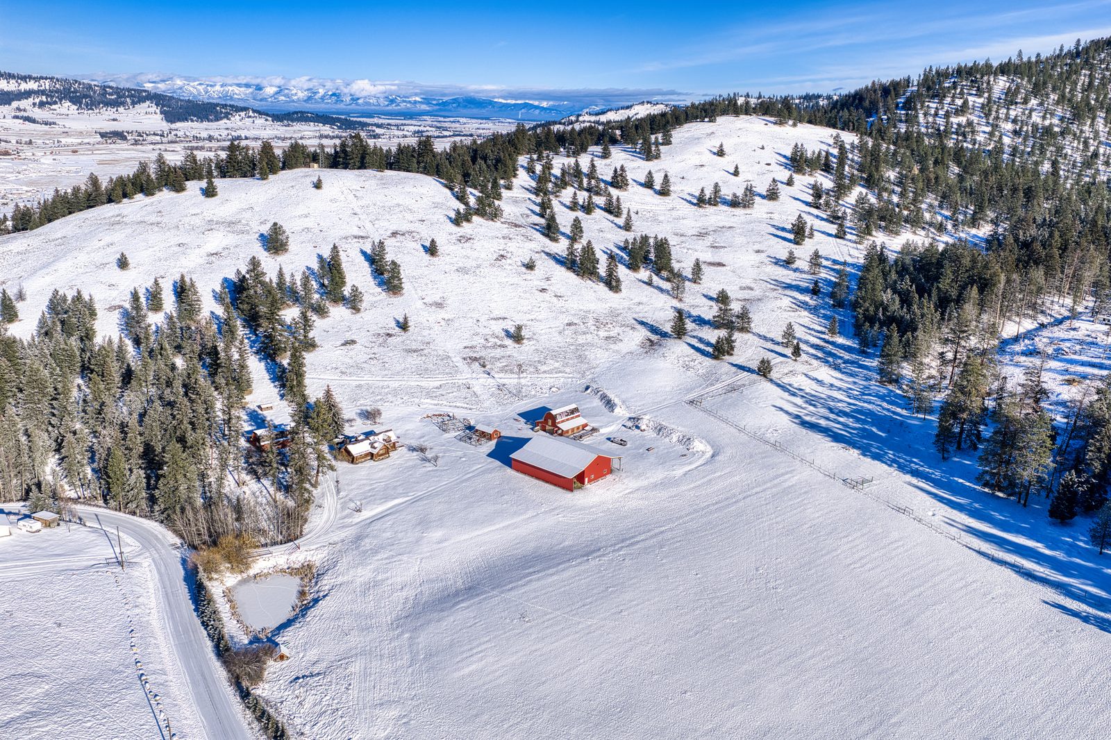 Aerial view of the 1675 Haywire Gulch homestead and red outbuildings under snow, with the Flathead Valley and distant mountains beyond